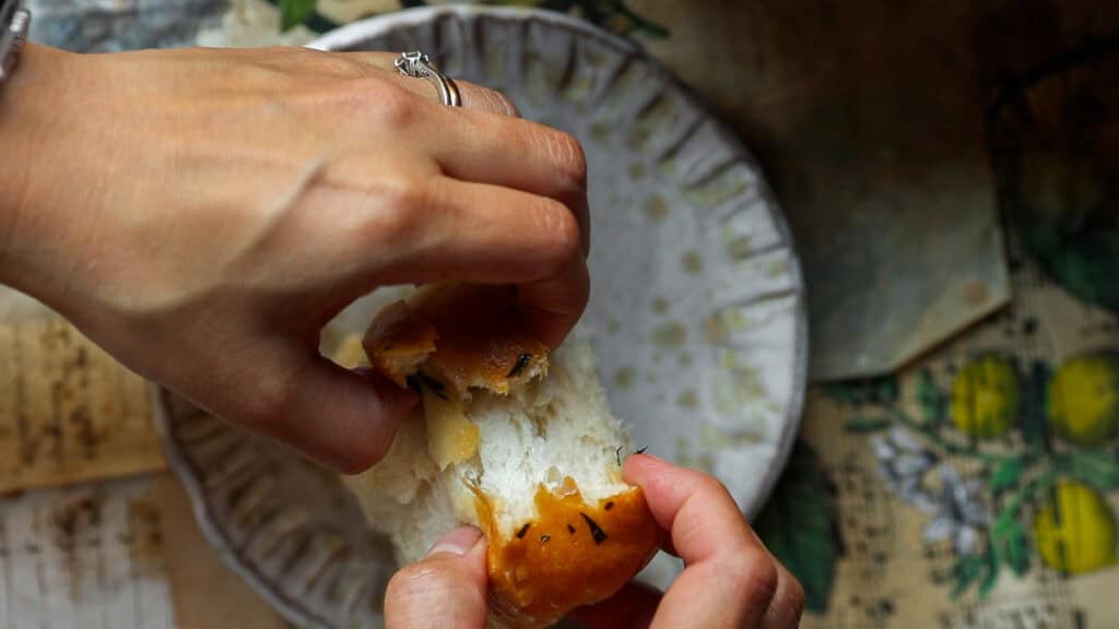 Two hands breaking a piece of bread above a decorative plate. The table underneath has a floral and lemon pattern. The person wears a ring on their right hand.